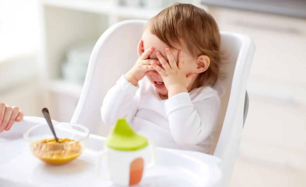 Mindful Living. Sustainable Living. Avoid Heavy Metals, Microplastics in Baby Food. Baby sitting in white high chair covers face with hands with puree in a glass bowl and a bottle in front of them.