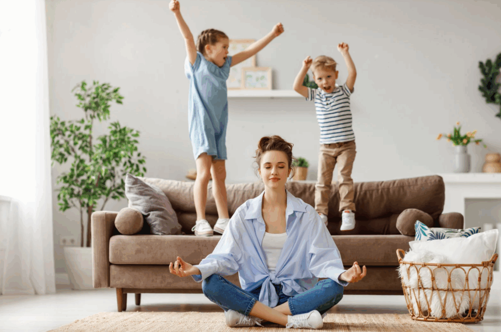 A mom sits in a meditative state on the floor of the living room next to a basket of laundry while her two children jump behind her on the couch.