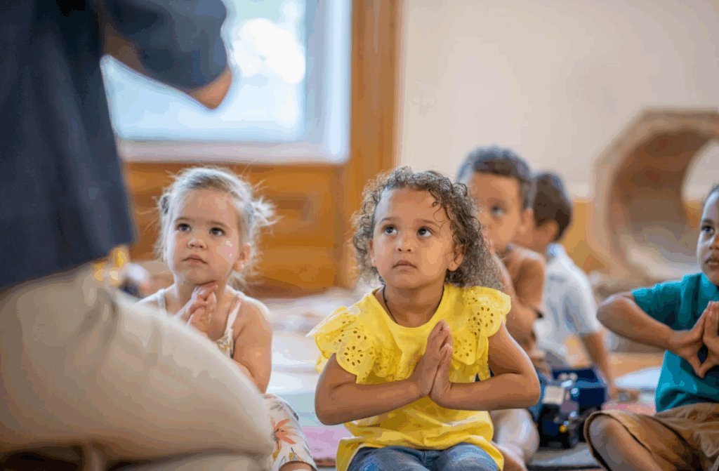 5 young children sitting on the floor their hands together looking to their teacher in a mindful moment.