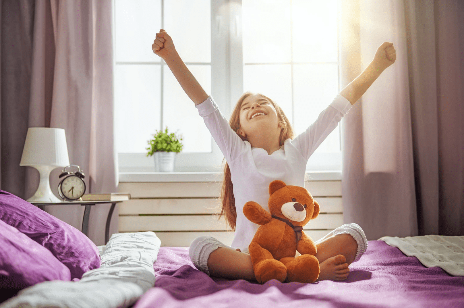 Young child stretching, arms up on a bed with her teddy bear in her lap, smiling.